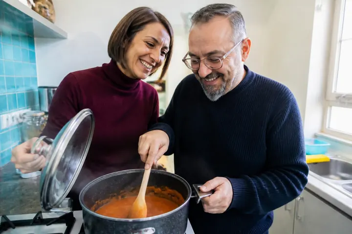 Küt kahve saçlı, bordo balıkçı yakalı bir kadın ile gözlüklü, keçi sakallı ve lacivert kazaklı bir adam küçük bir mutfakta tencerenin başında gülerek birlikte akşam yemeği hazırlıyor